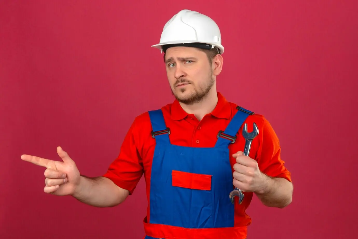 Männlicher Bauarbeiter in Arbeitskleidung und Schutzhelm hält einen Schraubenschlüssel, zeigt mit dem Finger zur Seite, blickt selbstbewusst und steht vor einer isolierten rosa Wand.